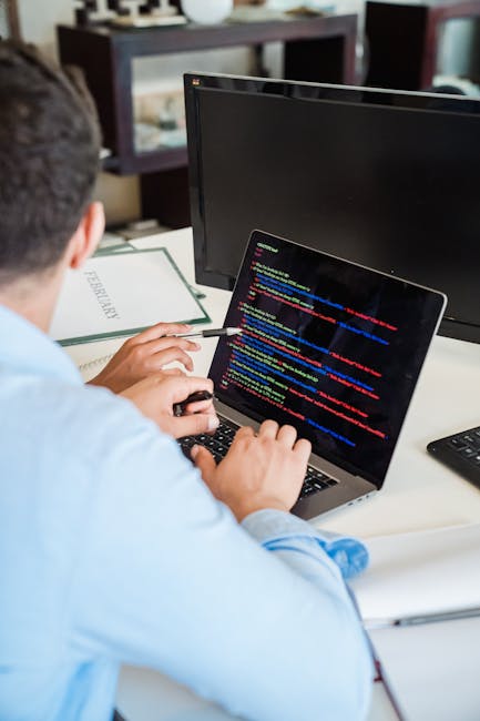 An individual working at a desk in a well-lit office space, focusing on a laptop displaying lines of colorful code. The desk holds a keyboard, a monitor, and a document labeled 'REFERENCES'. The backg