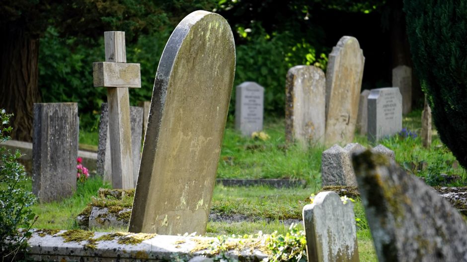 A small stone chapel with a peaked roof, pointed arch doorway, and decorative wooden details sits in the center of a cemetery. Surrounding the chapel, there are numerous white and grey gravestones arr