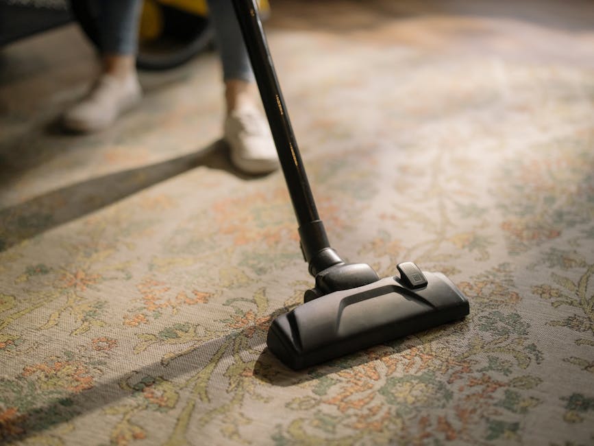 A person kneeling on a patterned Persian-style carpet while operating a yellow and black portable carpet cleaner from Brompton Carpet Cleaners. The individual is wearing a beige jacket and blue jeans,